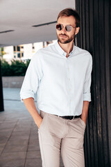 Fototapeta premium Handsome smiling stylish hipster lambersexual model. Modern man dressed in white shirt and trousers. Fashion male posing in the street urban background. Outdoors. In sunglasses