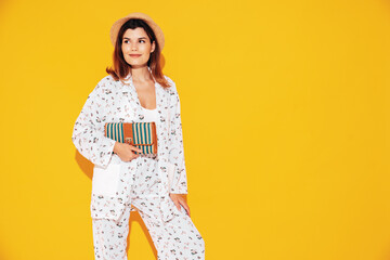 Obraz premium Young beautiful smiling female in trendy summer costume clothes. Carefree woman posing near yellow wall in studio. Positive model having fun indoors. Cheerful and happy. In hat. Holds clutch