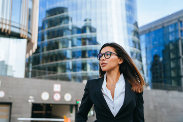 Purposeful European businesswoman in suit and glasses going to meeting looks forward with confident face expression. Brunette caucasian woman against skyscrapers. Businesspeople. Lawyer goes to meet