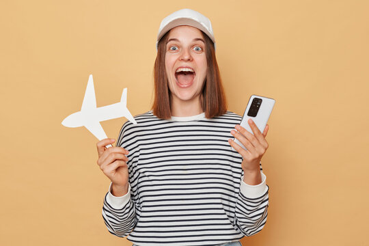 Excited Amazed Woman Wearing Striped Shirt And Baseball Cap Holding White Paper Plane And Using Smartphone Booking Tickets To Travel Abroad Standing Isolated  Over Beige Background