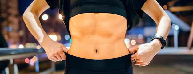 Unrecognizable young fit woman in black sportswear showing her muscular belly abs over a night city background
