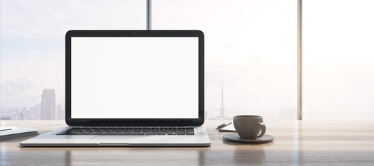 Fototapeta premium Wide image of laptop with empty white mock up monitor, coffee cup and notepad placed on wooden desktop. Window with bright city view in the background. 3D Rendering.