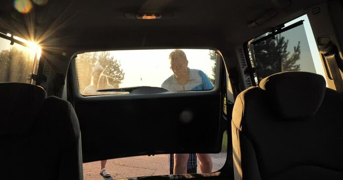 View from inside the car. A beautiful couple man and woman put their suitcases in the trunk and smile happily at each other. A cute couple of people are going on a trip by car and rejoice.