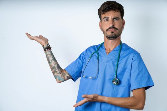 young caucasian doctor man wearing blue medical uniform pointing aside with both hands showing something strange and saying: I don't know what is this. Advertisement concept.