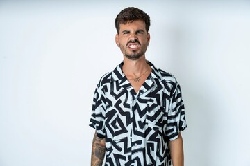 young caucasian man wearing printed shirt keeps teeth clenched, frowns face in dissatisfaction, irritated because of much duties.