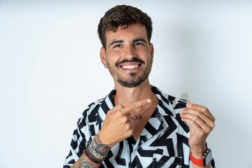young caucasian man wearing printed shirt holding an invisible aligner and pointing at it. Dental...