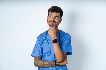 Thoughtful young caucasian doctor man wearing blue medical uniform holds chin and looks away...