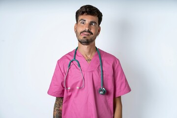 Funny young caucasian doctor man wearing pink medical uniform makes grimace and crosses eyes plays...