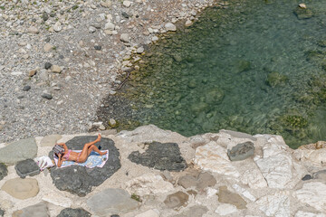 A woman sunbathes while reading a magazine lying on the rocks on the bank of the Trebbia river in Bobbio, Italy - obrazy, fototapety, plakaty