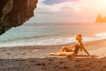  Woman travel sea. View of a woman in a black swimsuit from a sea cave Attractive woman enjoying the sea air sits on the beach and looks at the sea. Behind her are rocks and the sea © svetograph