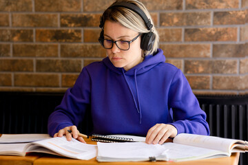 A girl is a student in large headphones at a table with notebooks and textbooks. A woman studies, writes a diploma and works with documents. Young woman in purple sweatshirt working with documents on