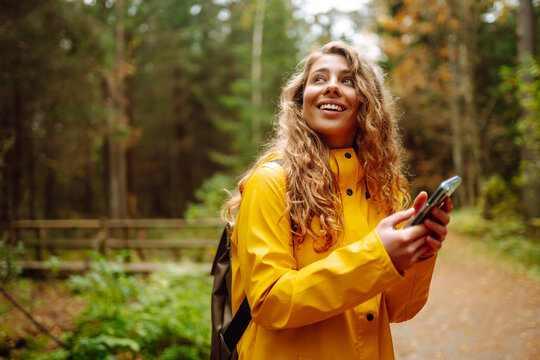 Young woman traveler in a yellow raincoat, holding a phone in her hands against the backdrop of a forest thicket. Smiling woman uses a mobile phone while walking in the forest.