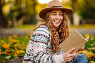 Beautiful woman in a hat and a stylish sweater sits on a mat in an autumn park and reads a book....