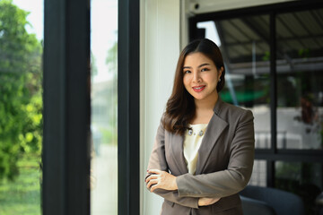 Portrait of successful millennial businesswoman in luxury suit standing with arms crossed and smiling at camera