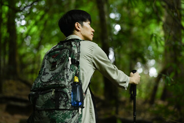 Rear view of man tourist with backpack and trekking sticks on a hiking trip in green forest