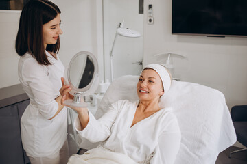 Aesthetic Procedure. Beautician Applying Clay Face Mask On Middle Aged Woman Face, Smiling Attractive Mature Lady Enjoying Anti-Aging Skincare Treatment In Cosmetology Studio, Above View