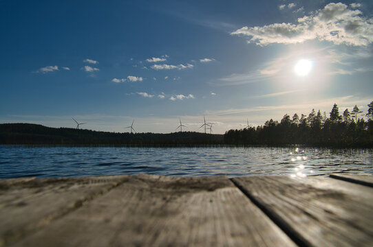 View over a footbridge into the blue water of a lake. Wind turbines in the background.