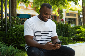 Fototapeta premium A young African-American man with a smartphone is sitting on a park bench