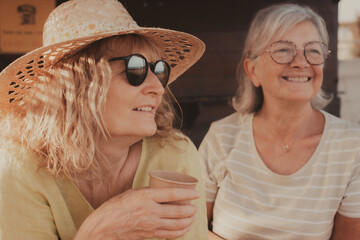 Smiling optimist couple of senior and middle age women with hats make confidences while drink a coffee cup. Two female friends enjoying freedom and relax in outdoors