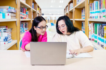 two female students at the desk using working on laptop with book shelf background