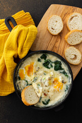Healthy breakfasts or dinners, fried eggs with cheese, spinach leaves and coconut milk in a cast-iron pan, sliced baguette on a cutting board on a brown background