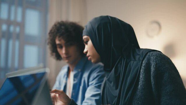 Young woman in hijab testing computer programs, talking with coworker in office