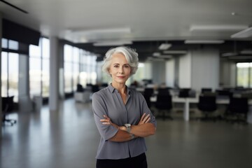 Elderly businesswoman looking at camera, standing with arms crossed in spacious modern office