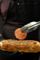 A man in a leather apron makes a hot dog, put grilled sausage in a bun, the concept of fast food and street food