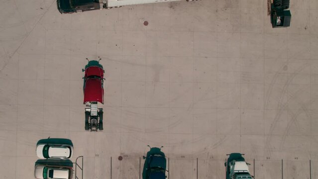 top down aerial of a red semi bobtail backing up to a reefer - dry van trailer to hook it up while another truck driver passing in front. Parking yard with other tractors and trailers parked