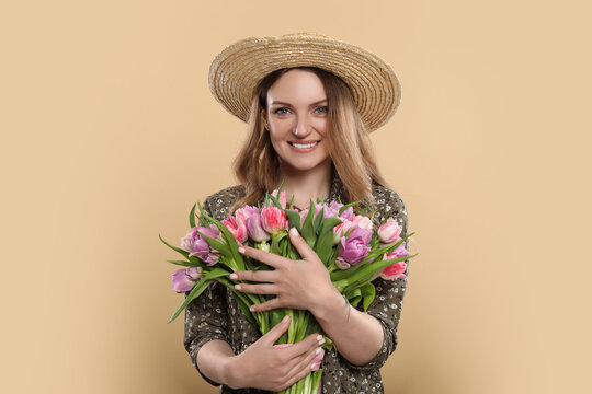 Happy young woman in straw hat holding bouquet of beautiful tulips on beige background