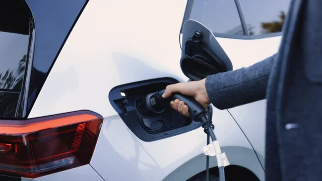 The driver of the electric car inserts the electrical connector to charge the batteries. Unrecognizable man attaching power cable to electric car. Electric vehicle Recharging battery charging port