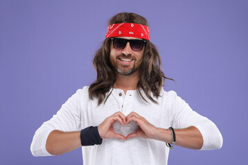 Stylish hippie man in sunglasses making heart with hands on violet background