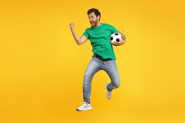 Emotional sports fan with soccer ball jumping on orange background