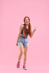 Stylish young hippie woman showing V-sign on pink background