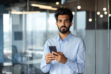 Portrait of young Indian male manager, office worker standing indoors and using phone. He looks seriously into the camera