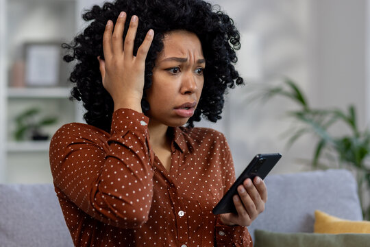 Worried African American woman sitting on sofa at home, looking at phone screen and holding head in shock