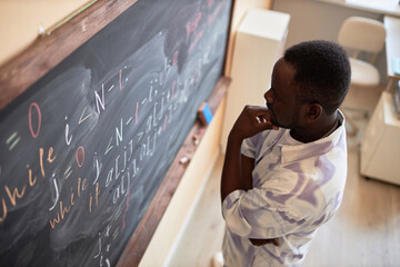 Pensive African American male student or teacher standing in front of blackboard with written...