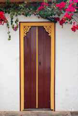 Old brown wooden double door with yellow ornaments on a white wall. Pink bougainvillea on top. Brazilian colonial old houses