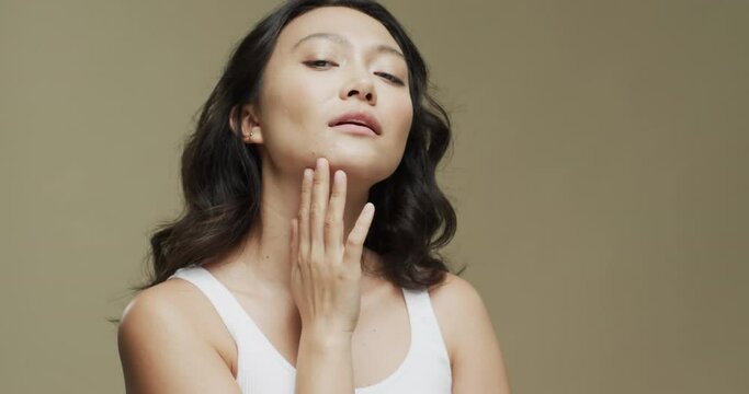 Asian woman with dark hair touching her face on beige background with copy space, slow motion