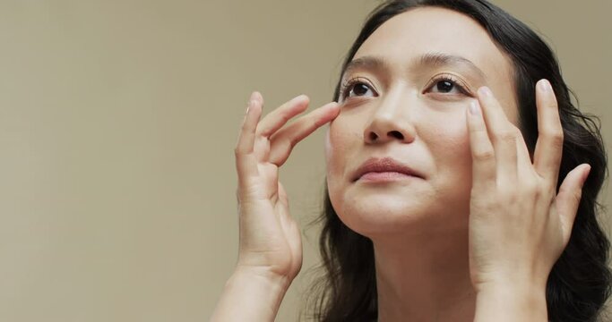 Asian woman with dark hair touching her face on beige background with copy space, slow motion