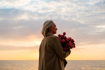 Carefree middle aged caucasian woman with bouquet of flowers on seashore, side view senior lady holding tulips on sea at sunset looking away