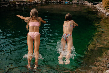 Two little girls jump in swimsuits into a lake with cold water on a hot summer day