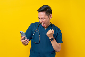 Excited young Asian male doctor or nurse wearing blue uniform using mobile phone and reading news while celebrating success isolated on yellow background. Healthcare medicine concept