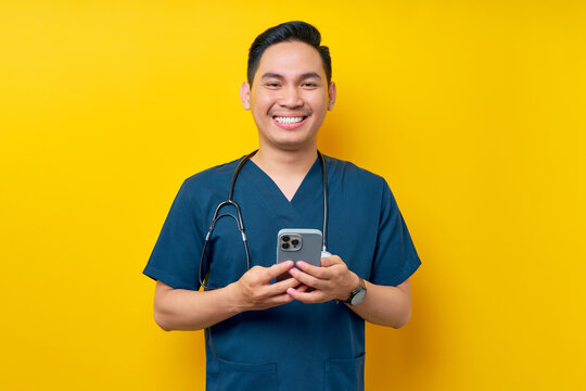 Smiling Happy Young Asian Male Doctor Or Nurse Wearing Blue Uniform Holding Mobile Phone And Looking At A Camera Isolated On Yellow Background. Healthcare Medicine Concept