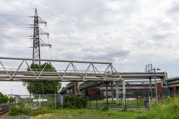Industrial area with aerial pipelines and electric masts. The sky is covered with gray clouds.