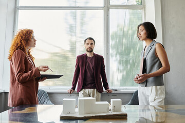 redhead designer talking to businesswoman near investor in office, building model presentation