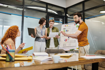 bearded businessman pointing at document near colleagues in meeting room with graphs on flip chart