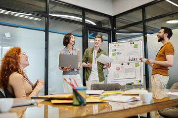excited team laughing near flip chart with graphs in meeting room, successful collaboration