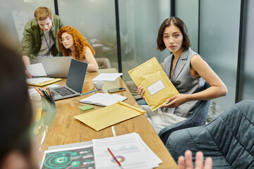 young businesswoman with envelope near colleagues talking in meeting room, laptops, graphs