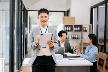 Young attractive Asian male office worker business suits smiling at camera with working notepad, tablet and laptop documents in office